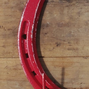 May include: A red horseshoe with white paint accents. The horseshoe is hanging on a wooden surface.