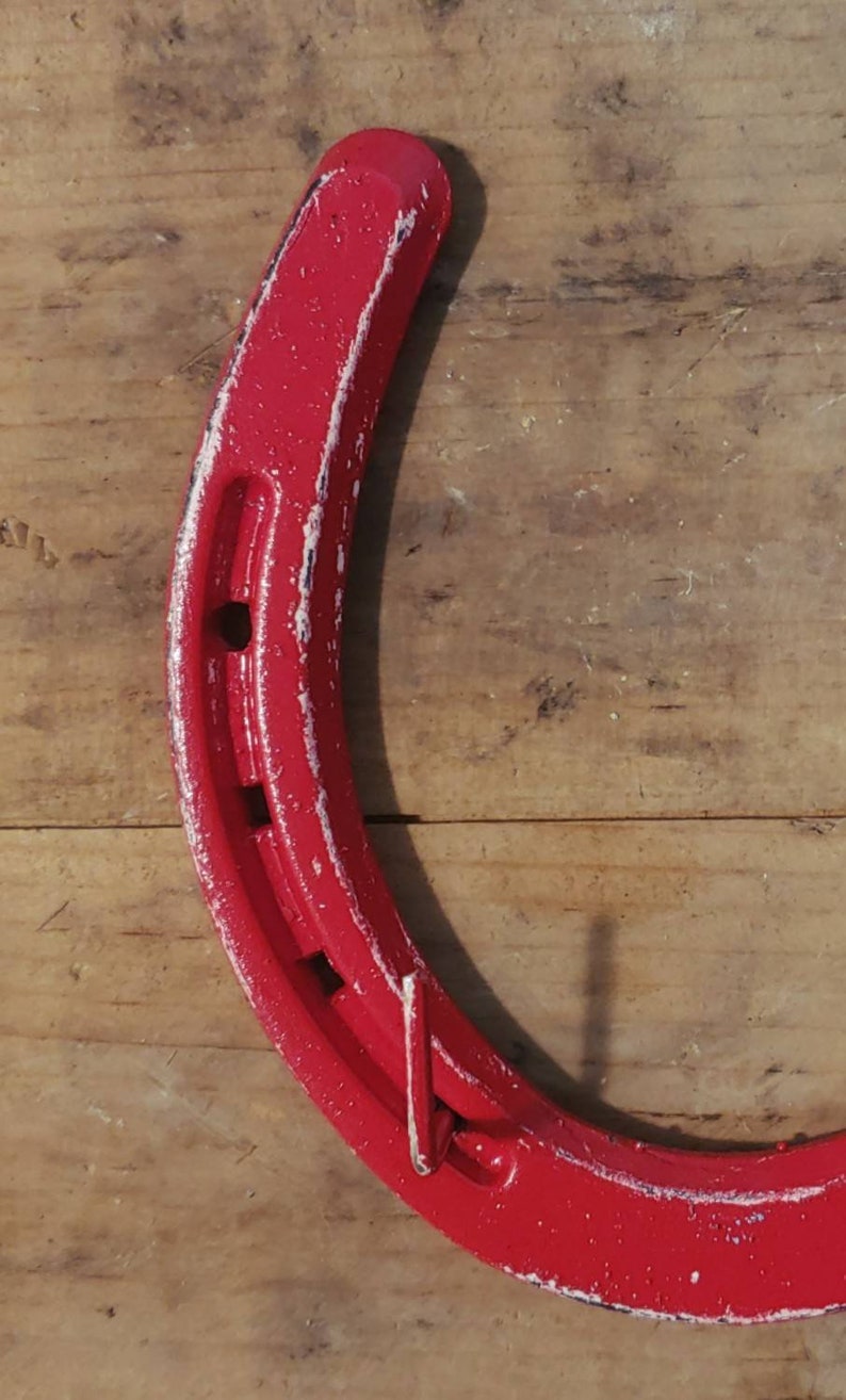 May include: A red horseshoe with white paint accents. The horseshoe is hanging on a wooden surface.