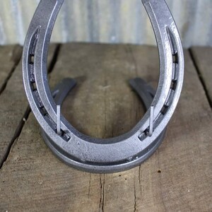May include: A silver horseshoe with two metal hooks attached to the top. The horseshoe is sitting on a wooden surface.