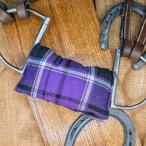 Op de afbeelding: Een paars en zwart geruit paardenbit met een zilveren metalen frame. Het bit ligt op een houten oppervlak met een bruine jute achtergrond.