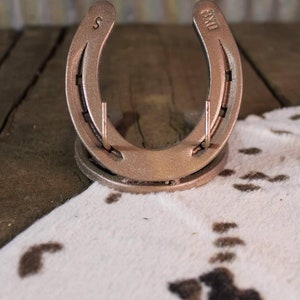 May include: A gold-colored horseshoe with the number "8" and the letter "S" engraved on it. The horseshoe is on a white and brown cowhide rug.