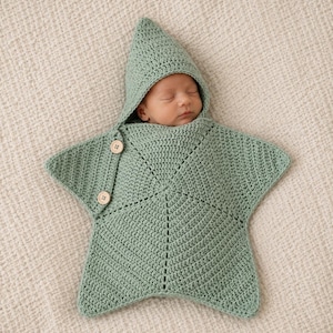 May include: A sage green crocheted star-shaped swaddle blanket with a pointed hood. The garment features two light-colored wooden buttons on the side flap and is designed to wrap around an infant, leaving only the face visible within the hood.
