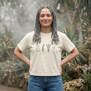 May include: A cream-colored short-sleeve t-shirt featuring a black line-art graphic on the chest. The design consists of a salt shaker, a sprig of rosemary, a bundle of lavender, and a small heart symbol arranged in a horizontal row.