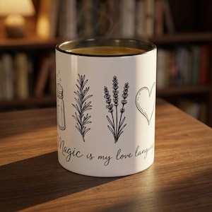 May include: A white ceramic mug with a black rim, featuring black line art illustrations of a salt shaker, a sprig of rosemary, a sprig of lavender, and a heart. Below the illustrations, the text reads, Magic is my love language.