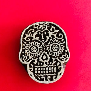May include: A wooden skull stamp with intricate floral and swirl designs. The eyes are sunflower-shaped, and the teeth are clearly defined. The stamp is dark brown with a light brown design, set against a bright red background.