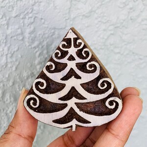 Può includere: Un timbro triangolare in legno con un disegno di albero di Natale. Il timbro è marrone con un motivo ad albero bianco stilizzato con dettagli a spirale. Il timbro è tenuto in mano su uno sfondo strutturato di colore chiaro.