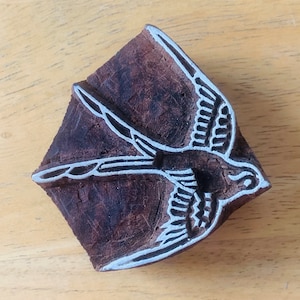 May include: A wooden block print with a carved design of a bird in flight. The bird is depicted with outstretched wings and detailed feathers. The block is a dark brown color with white accents.