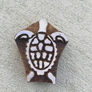 Puede incluir: Un bloque de madera con un diseño de tortuga. La tortuga es blanca y el bloque es marrón. El bloque tiene forma de diamante con una parte posterior plana. El caparazón de la tortuga tiene un patrón detallado.