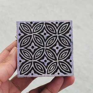 Può includere: Un blocco di legno quadrato con un motivo floreale ripetuto. Il blocco è dipinto di un colore viola chiaro, con il motivo floreale in marrone scuro. Il disegno presenta fiori a quattro petali e piccoli punti. Il blocco è tenuto in una mano.
