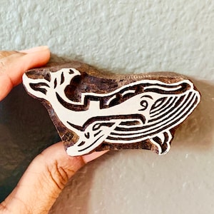 Op de afbeelding: Een houtsnijwerk van een walvis met ingewikkelde details. De walvis is gesneden in wit tegen een donkerbruine houten achtergrond.