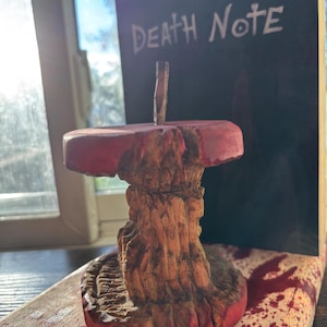 May include: A carved wooden apple core sculpture with a red painted top and base. The sculpture is in front of a black book with the words "DEATH NOTE" written in white. The base it sits on has red splatters.