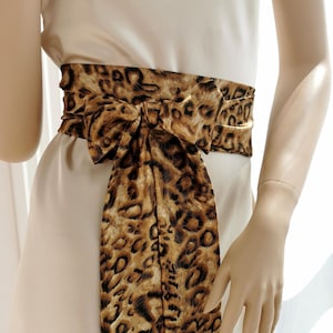 May include: A brown and black leopard print satin sash with a bow tied in the front. The sash is tied around a white dress.