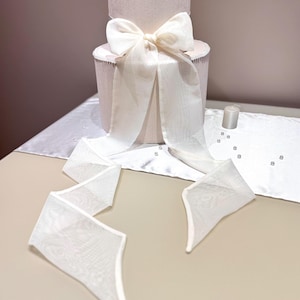 Puede incluir: Una caja de regalo cilíndrica rosa claro de dos niveles, con un gran lazo blanco y una cinta blanca. La parte superior de la caja está adornada con una flor blanca. La caja está sobre un camino de mesa blanco.