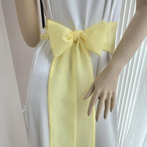 May include: A white dress with a large, pale yellow bow tied at the waist. The bow is made of a sheer fabric and hangs down the front of the dress. The dress is simple and elegant, with a flowing silhouette.