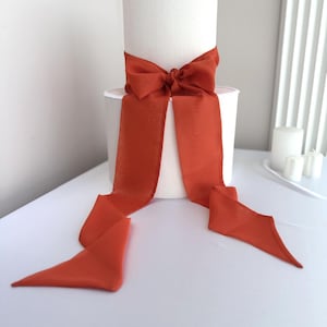 Puede incluir: Un soporte para pasteles cilíndrico blanco con un borde blanco decorativo en la parte superior. Una gran cinta de color naranja quemado está atada alrededor del soporte en forma de lazo, con largas colas.