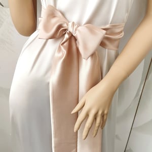 May include: A blush pink satin ribbon tied in a bow around a white dress. The ribbon is long and flowing, with the ends trailing down the sides of the dress.