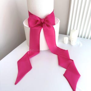 May include: A white, cylindrical lampshade adorned with a vibrant pink bow. The bow's long ribbons cascade downwards, offering a bold contrast against the white. The lampshade is positioned on a white surface, creating a clean aesthetic.