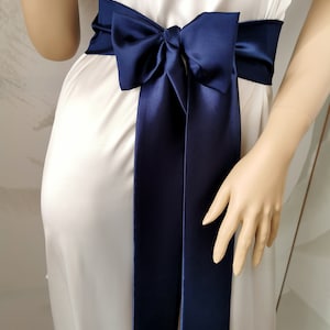 May include: A navy blue satin sash with a bow tied in the center. The sash is tied around a white dress.