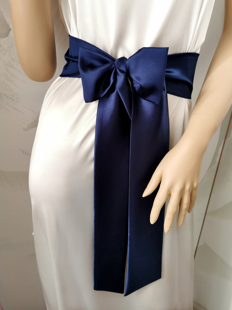May include: A navy blue satin sash with a bow tied in the center. The sash is tied around a white dress.