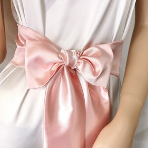 May include: A blush pink satin ribbon tied in a bow around a white dress.