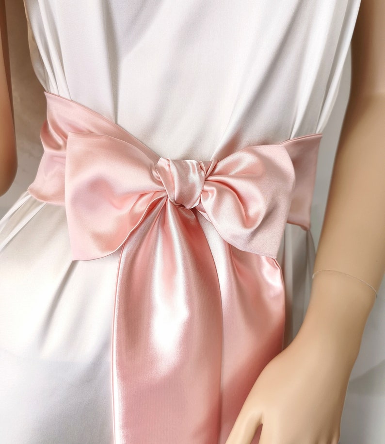 May include: A blush pink satin ribbon tied in a bow around a white dress.