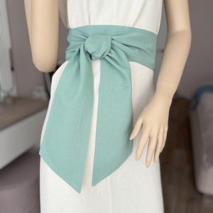 Op de afbeelding: Een saliegroene linnen band geknoopt om de taille van een witte linnen jurk. De band is aan de voorkant in een strik geknoopt.