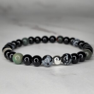 May include: A black and green beaded bracelet with a silver bead. The bracelet is made with black onyx, green aventurine, and snowflake obsidian beads.