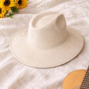 May include: A cream-coloured fedora hat with a wide brim, resting on a textured, off-white surface. The hat has a simple band around the crown. A bouquet of yellow sunflowers and a wooden acoustic guitar are also in the image.