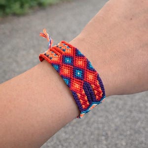 May include: A colorful woven friendship bracelet with a geometric pattern. The bracelet features a repeating diamond design in shades of orange, red, blue, and purple. The bracelet is handmade and has a braided closure.