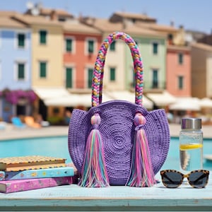 May include: A purple woven handbag with a braided handle and colorful tassels, next to a water bottle, sunglasses, and books. The bag is round and has a circular woven design. The sunglasses have a tortoiseshell pattern.