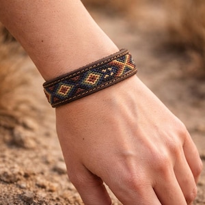May include: A brown leather bracelet featuring a central woven textile band with a geometric diamond pattern in shades of navy blue, olive green, and mustard yellow, secured with visible stitching along the top and bottom edges of the band.
