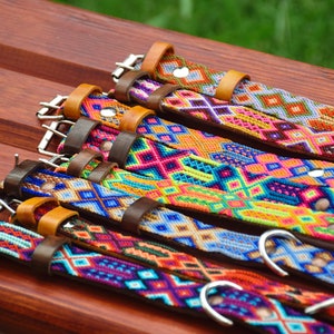 May include: A collection of five dog collars featuring intricate woven geometric patterns in vibrant colors including orange, blue, pink, and green. Each collar is constructed with a brown leather base, a metal buckle, and a metal D-ring attachment.