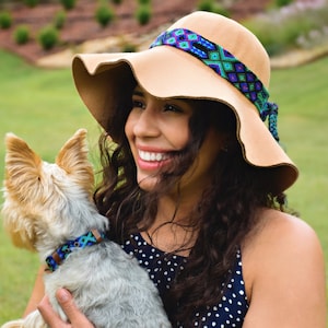 May include: A woman with long brown hair wearing a wide-brimmed tan hat with a colorful woven band is holding a small, gray and brown dog with a blue and green patterned collar.