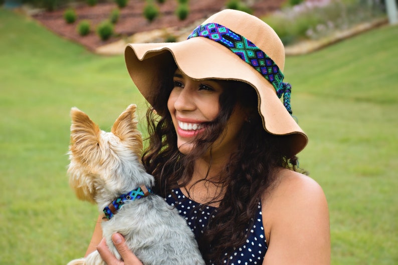 May include: A woman with long brown hair wearing a wide-brimmed tan hat with a colorful woven band is holding a small, gray and brown dog with a blue and green patterned collar.