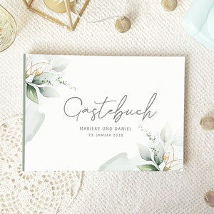 May include: A white guest book with a watercolour design of green leaves and gold accents. The text "Gästebuch" is written in cursive script. The text "Marieke und Daniel" and "22. Januar 2023" is printed below the title.