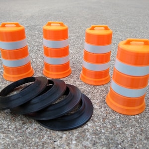 May include: Four orange and white traffic cones with black rubber rings. The cones are arranged in a row on a gray concrete surface.