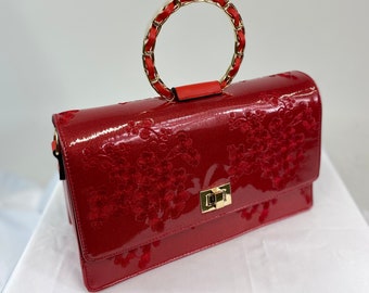 Può includere: Una borsa a mano in pelle verniciata rossa con un manico superiore a catena dorata e ricamo floreale. La borsa ha una chiusura a lembo con una fibbia dorata.