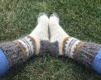 May include: A pair of white, grey, and yellow wool socks with a fluffy texture. The socks are being worn on a person's feet and are resting on a patch of green grass.