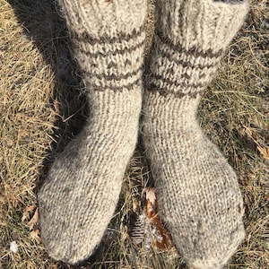 May include: A pair of thick, hand-knitted wool socks with brown stripes. The socks are made of a natural, light brown wool and are shown on a person's feet against a background of dry grass.