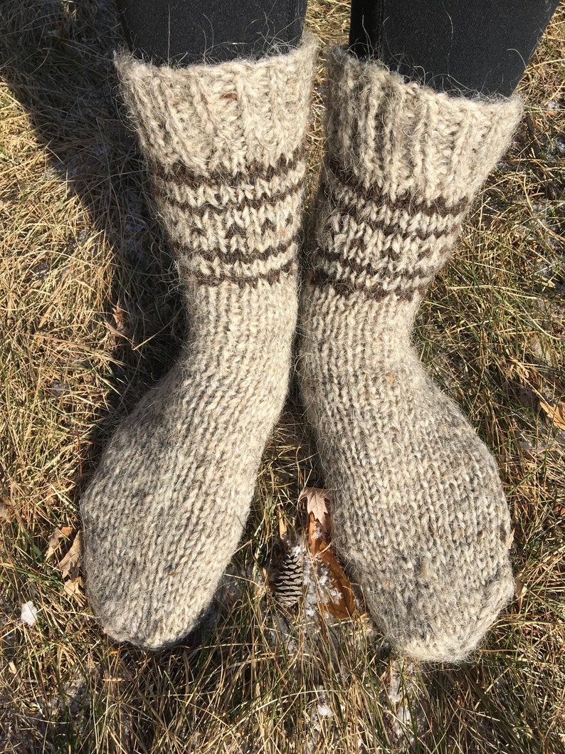 May include: A pair of thick, hand-knitted wool socks with brown stripes. The socks are made of a natural, light brown wool and are shown on a person's feet against a background of dry grass.