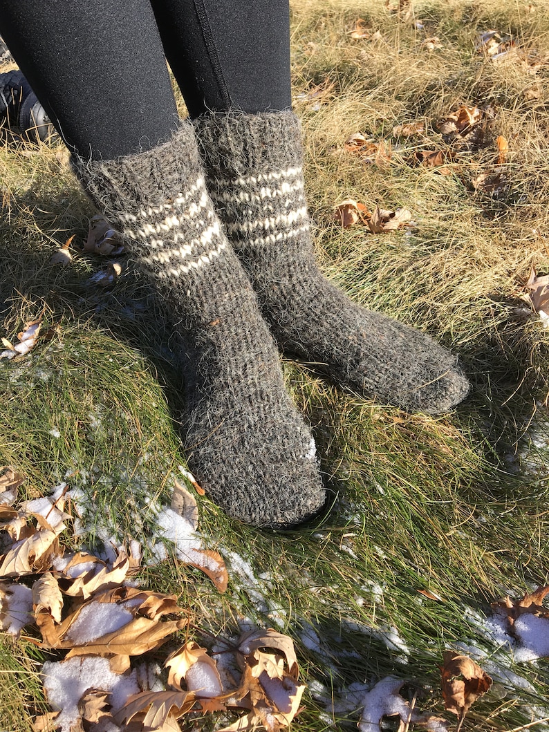 May include: A pair of gray wool socks with white stripes. The socks are worn and are sitting on a bed of green grass with brown leaves.