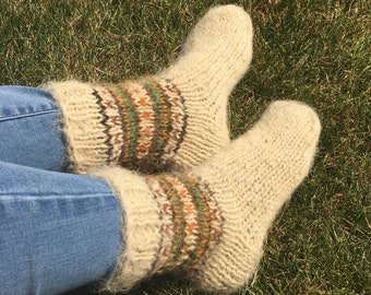 May include: A pair of white wool socks with brown, orange, green and blue knitted patterns. The socks are worn on a person's feet and are lying on green grass.