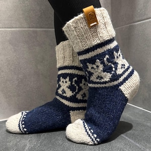 May include: A pair of knitted socks in navy blue and cream. The socks feature a patterned design in cream on the navy blue sections. The cuffs are cream with a brown leather tag. The socks are made of a thick, warm material.