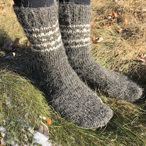 May include: A pair of gray wool socks with white stripes. The socks are thick and fuzzy, and they are worn by a person standing on a bed of green grass.