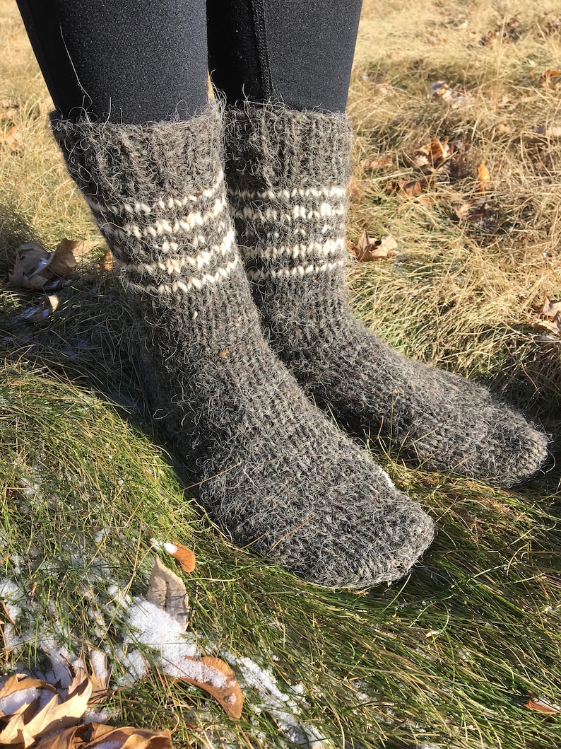 May include: A pair of gray wool socks with white stripes. The socks are thick and fuzzy, and they are worn by a person standing on a bed of green grass.