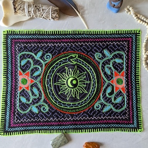 以下が含まれることがあります： ネオンの緑色の縁取りと複雑な刺繍が施された長方形の黒い織物。デザインは、ターコイズ、ピンク、オレンジの花柄と幾何学模様に囲まれた、太陽と蛇の中央モチーフを特徴としています。その他のアイテムには、羽、ビーズ、小さな置物などがあります。