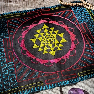 May include: Black embroidered textile with a central yellow geometric design, encircled by a pink floral pattern. The border features blue, red, and yellow stitching. A small purple crystal is visible.