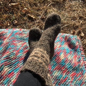 May include: A pair of brown wool socks with white stripes, worn on a person's feet. The socks are resting on a colorful striped blanket.