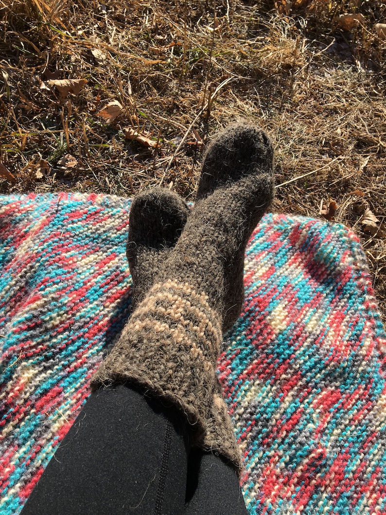 May include: A pair of brown wool socks with white stripes, worn on a person's feet. The socks are resting on a colorful striped blanket.