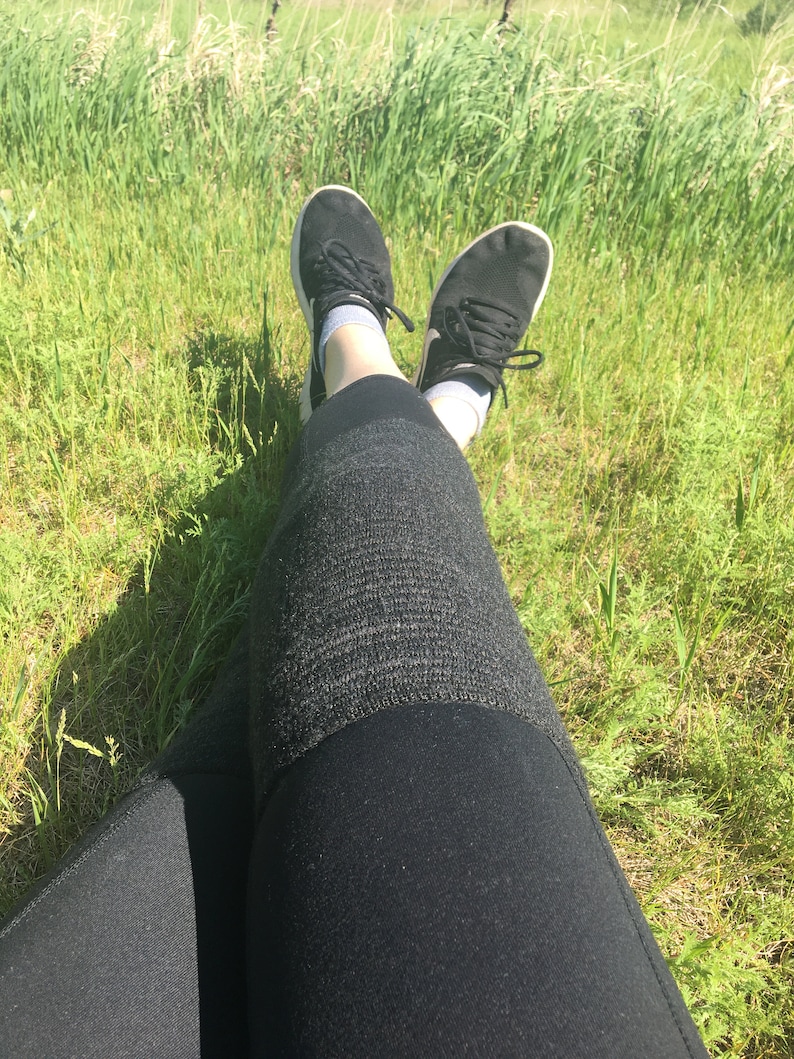 May include: A person wearing black leggings and black sneakers with white soles, sitting in a grassy field.